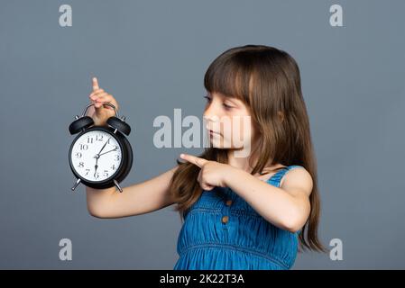 Une petite fille d'avant-chooler tient un réveil rond dans ses mains et montre l'heure avec son index. Isolé sur un fond gris. Il est temps d'étudier et de ne pas être en retard pour l'école. Photo de haute qualité Banque D'Images