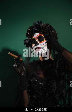 Femme dans le jour mexicain de maquillage mort et costume tenant le cigare sur fond vert, image de stock Banque D'Images