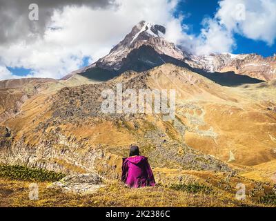 Thoughtful woman sit on grass admire beautiful KAzbek mountain snowy peak in autumn. Hike to gergeti glacier route solo. Travel in caucasus concept Stock Photo