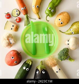 Backgound with attractive plate of funny fruits and vegetables served for child with green children's cutlery on white table. Top view. Stock Photo