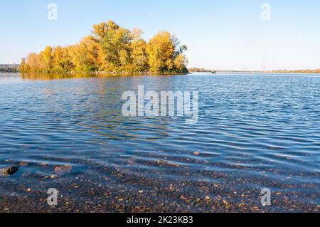 Sunny fall day close to the blue Dnieper river in Kiev, Ukraine, with autumn trees around. Stones appear under the clear water. Stock Photo