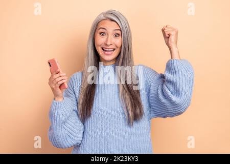 Photo de joyeuse joyeuse femme drôle de lever les poings dans la victoire voir de grandes affaires dans le téléphone isolé sur fond beige couleur Banque D'Images