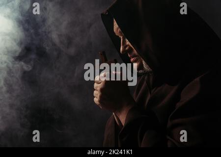 vue latérale du moine en robe tenant la croix sainte tout en priant sur le noir avec de la fumée, image de stock Banque D'Images