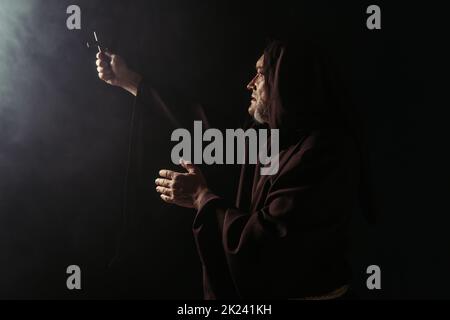 monk en robe à capuchon noire tenant saint crucifix dans la main tendue sur fond sombre, image de stock Banque D'Images