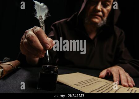 moine flou tenant plume près de l'inkpot et manuscrit ancien isolé sur noir, image de stock Banque D'Images