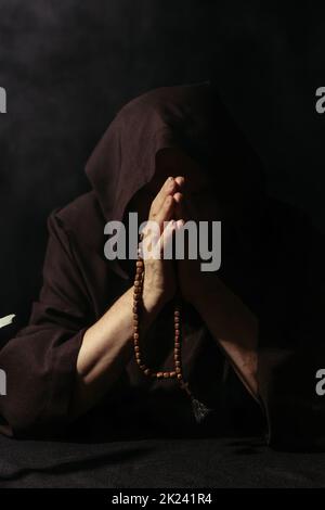 moine anonyme en robe à capuchon priant avec des perles rosaires isolées sur noir, image de stock Banque D'Images