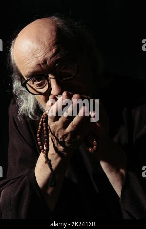 moine senior dans les lunettes tenant le rosaire et regardant la caméra isolée sur noir, image de stock Banque D'Images