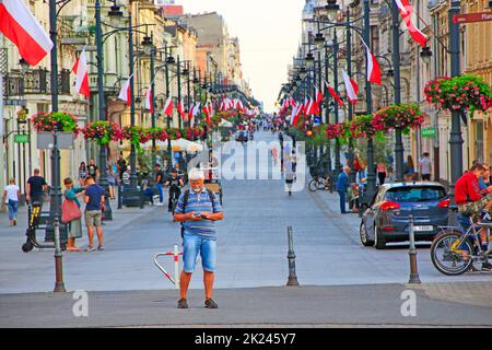 Homme avec téléphone mobile debout dans la rue centrale de Lodz. Rue centrale de Lodz décorée avec des drapeaux polonais. Personnes marchant le long de la rue de la ville Piotrko Banque D'Images