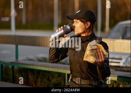 Une femme de police affamée mangeant un sandwich assis sur le banc du parc. La COP femelle en uniforme fait une pause Banque D'Images