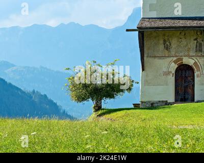 Un pré d'été avec un ciel bleu et quelques nuages blancs avec une grande chaîne de montagnes en arrière-plan.Une petite chapelle avec un arbre fruitier peut être Banque D'Images