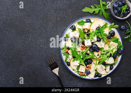 Delicious arugula salad with pears, blueberries, roquefort cheese and walnuts. Black kitchen table background, top view Banque D'Images