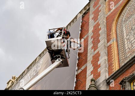 Amsterdam, Netherlands - August 2022: People standing on a cherry picker platform fixing a large banner to the outside wall of the famous Rijks Museum Stock Photo