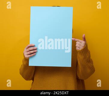 femme dans un chandail orange tient une feuille de papier vierge sur un fond jaune.Place pour une inscription, une publicité, des informations Banque D'Images