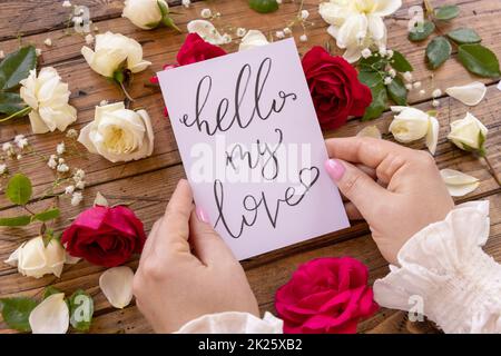 Mains avec carte BONJOUR MON AMOUR près de fleurs rouges et crème gros plan sur une table en bois Banque D'Images