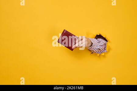 main tient un portefeuille en cuir marron sur un fond jaune, une partie du corps dépasse d'un trou déchiré dans le papier Banque D'Images