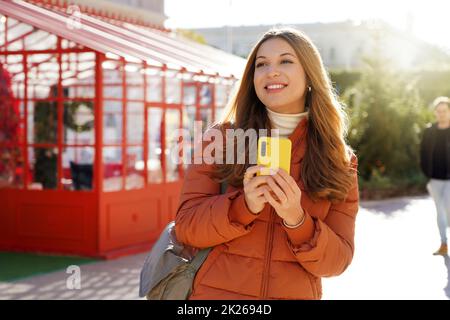 Décontracté belle femme portant un manteau tenant un téléphone mobile regardant devant marcher dehors Banque D'Images