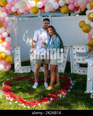 Newly engaged couple standing in a heart made of red and white rose pedels with letters spelling love behind them. Banque D'Images
