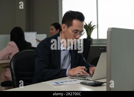 Homme d'affaires travaillant sur un ordinateur portable, assis au bureau de son poste de travail avec des collègues en arrière-plan. Banque D'Images