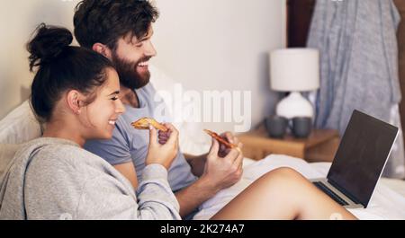 Date nuit fait correctement.Photo d'un jeune couple heureux en utilisant un ordinateur portable et en mangeant de la pizza tout en se relaxant sur le lit à la maison. Banque D'Images