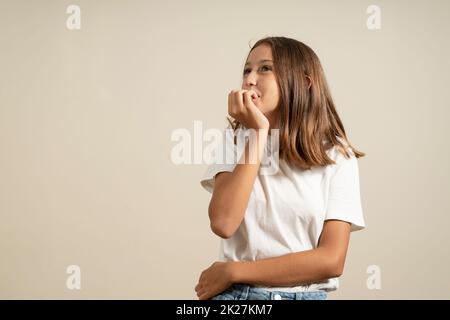 Close up portrait of a worried teenage girl in white t-shirt biting her nails isolated over beige background. Stock Photo