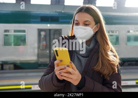 Jeune femme avec masque médical acheter un billet en ligne avec smartphone à la gare Banque D'Images