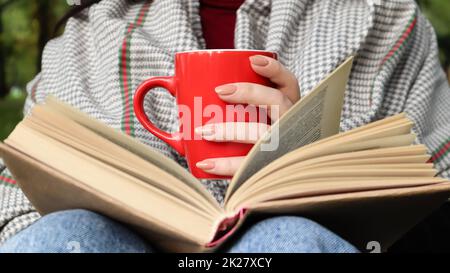 Une fille vêtue d'un manteau et d'un foulard dans la forêt d'automne tient un livre et une tasse avec une boisson chaude dans ses mains en gros plan dans un parc de la ville lors d'une journée chaude. Le concept de lecture, de détente et de confort. Banque D'Images