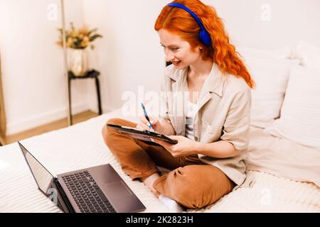 Hipster teen girl school student with orange hair wear headphone write notes watch video online webinar learn on laptop sit in bed distance elearning Stock Photo