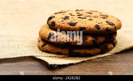 Biscuits aux pépites de chocolat américain sur une table en bois marron et sur une serviette en lin en gros plan. Pâte croquante traditionnelle arrondie avec copeaux de chocolat. Boulangerie. Délicieux dessert, pâtisseries. La vie rurale encore. Banque D'Images