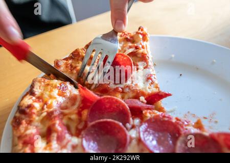 Les mains de garçon ont coupé la pizza de salami en morceaux avec fourchette et couteau sur la plaque à pizza en gros plan macro avec le fromage délicieux et le salami comme graisse alimentaire rapide malsaine pour les adolescents affamés régime italien de délicatesse Banque D'Images