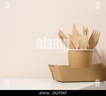 assiettes et tasses en carton brun, fourchettes et couteaux en bois sur une table blanche, fond beige.Vaisselle écologique, sans gaspillage Banque D'Images