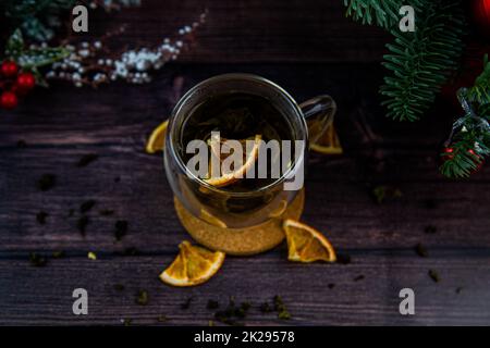 Une tasse de thé transparente est placée sur une table sombre, sur un support en bois, des tranches d'orange séchées flottent dans la tasse, des tranches d'agrumes se trouvent à côté, du thé est dispersé, une photo dans une touche sombre Banque D'Images
