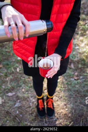 Une fille dans un gilet orange vif verse le thé chaud d'un thermos, une collation en plein air.Voyage, trekking et randonnée. Banque D'Images