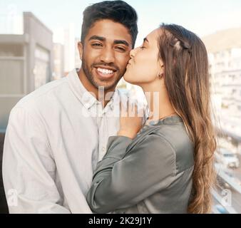 Couple heureux, amour et baiser sur la joue de la petite amie liant avec le petit ami debout sur un balcon de bâtiment de ville pendant des vacances. Portrait de l'indien Banque D'Images