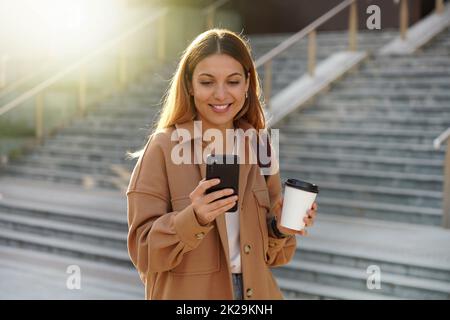 Jeune femme blonde enchanteresse qui attend un message téléphonique tout en portant une tasse de café dans la rue. Fille élégante vêque de vêtements de printemps pour smartphone et cappuccino à emporter. Banque D'Images