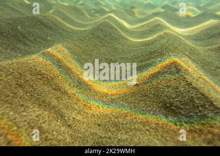 Photo sous-marine - La lumière réfractée sur la surface de la mer formant un arc-en-ciel sur les 'dunes' de sable en eau peu profonde près de la plage. Résumé fond marin. Banque D'Images