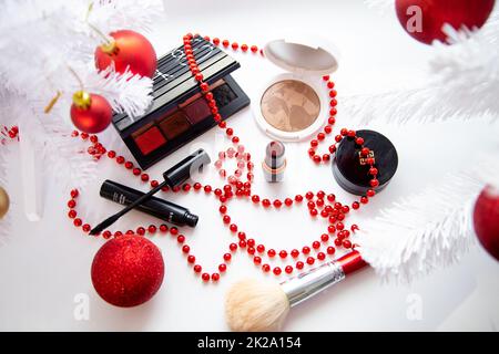 Poudre et bronzant, ombre à paupières, rouge à lèvres, blush et un pinceau de différentes marques se trouvent sur un fond blanc entouré de branches blanches moelleuses d'un arbre de Noël et de boules rouges de Noël. Banque D'Images