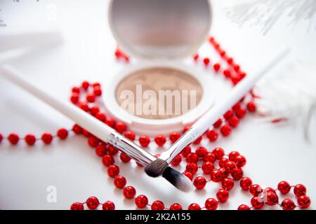 Un pilbox blanc à la poudre et à la poudre bronzante se trouve sur un fond blanc, de fines pinceaux de maquillage et des perles rouges brillantes se trouvent à l'avant. Banque D'Images