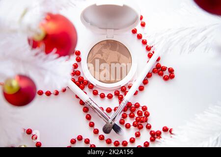 Un coussin blanc avec poudre et bronze se trouve sur un fond blanc parmi un arbre de Noël blanc moelleux avec des jouets rouges, de fines pinceaux de maquillage et des perles rouges brillantes se trouvent devant. Banque D'Images