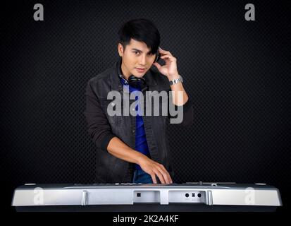 Jeune homme asiatique avec casque jouant un clavier électrique devant un mur insonorisant noir. Musicien produisant de la musique dans un studio d'enregistrement professionnel. Banque D'Images