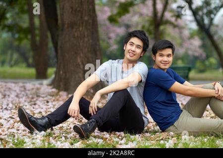 C'est toujours là pour les uns les autres. Mignon jeune gay asiatique couple souriant ensemble tout en étant assis dans le parc. Banque D'Images