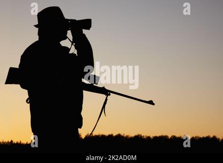 Suivi de la faune. Une silhouette d'un garde-corps tenant son fusil et regardant à travers ses jumelles au soleil se lève à l'extérieur. Banque D'Images