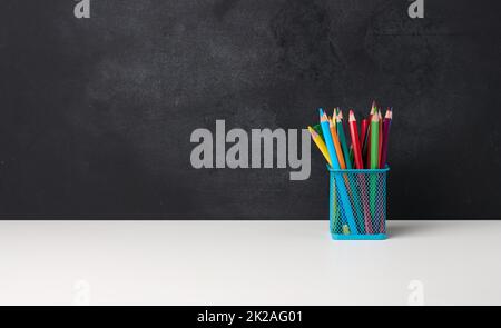 verre métallique avec stylos, crayons et stylos-feutres et ciseaux sur le fond d'un tableau noir vide de craie, table blanche Banque D'Images