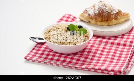 Flocons d'avoine crus dans une assiette en céramique blanche et croissant sur une table blanche, petit déjeuner Banque D'Images