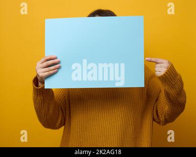 femme dans un chandail orange tient une feuille de papier vierge sur un fond jaune.Place pour une inscription, une publicité, des informations Banque D'Images