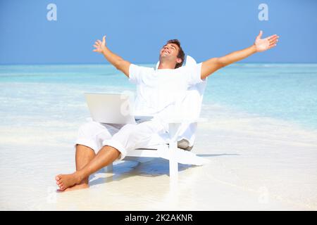 Loin de la vie trépidante. Portrait d'un homme insouciant qui dépasse ses bras avec un ordinateur portable sur la plage. Banque D'Images