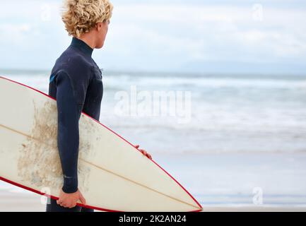 À la recherche de cette vague parfaite Un jeune surfeur debout sur la plage avec l'océan en arrière-plan. Banque D'Images