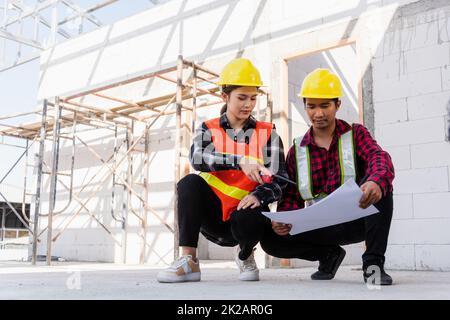 L'architecte et le client discutent de l'aide à la création d'un plan avec le plan du bâtiment sur le chantier de construction. Un homme et une femme de travail de contremaître d'ingénieur asiatique se rencontrent pour discuter du projet de papier à dessin, travail d'équipe Banque D'Images