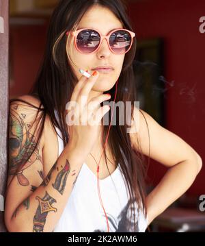 Rebel à cœur. Photo d'une belle jeune femme tatouée fumant une cigarette tout en écoutant de la musique. Banque D'Images