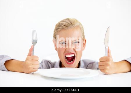 C’est ridicule et affamé. Une jeune femme furieuse assise devant une assiette vide tout en tenant son couteau et sa fourchette. Banque D'Images