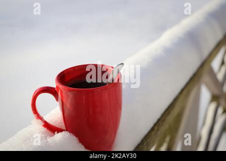 Tasse de café chaud ou de thé à la vapeur debout sur la table extérieure dans le matin d'hiver de neige. Confortable mug rouge festif avec boisson chaude dans le jardin d'hiver. Concept du matin de Noël Banque D'Images